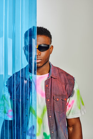 Portrait of fashionable young afroamerican man in sunglasses, denim vest and t-shirt standing behind...