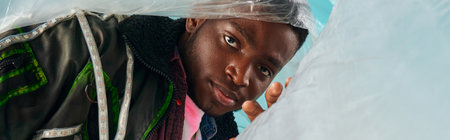 Portrait of stylish african american young man in outwear jacket with led stripes posing with glossy cellophane on turquoise background, urban outfit and modern pose, banner, creative expressionの写真素材