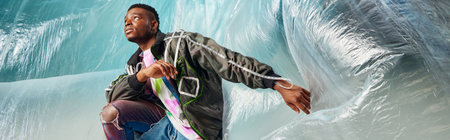 Young afroamerican man in modern outwear jacket and ripped jeans looking away near cellophane on...