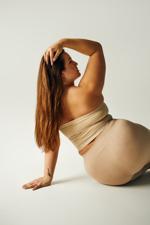 back view of charming woman with plus size body with strapless top, bare shoulders and underwear touching hair while sitting in studio on grey background, body positive, tattoo translation: harmonyの写真素材