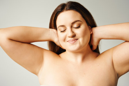 portrait of pleased woman with plus size body and closed eyes touching hair and posing with bare shoulders isolated on grey background in studio, body positive, self-loveの写真素材
