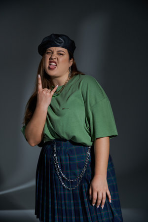stylish plus size woman posing in leather beret, green t-shirt and plaid skirt looking at camera,...