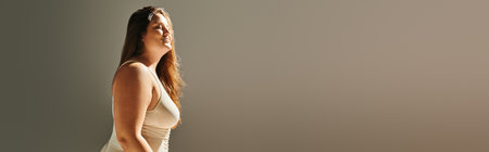pleased plus size woman with long hair wearing beige bodysuit and enjoying bright lighting while posing on grey background with studio light, body positive, figure type, diversity of body, bannerの写真素材