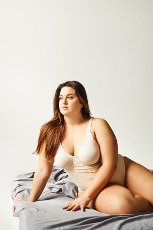 brunette plus size woman with natural makeup and long hair wearing beige bodysuit and posing on bed with grey bedding while looking away, body positive, figure type, caucasianの写真素材