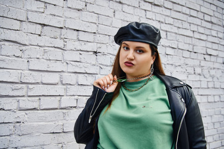 stylish plus size woman in leather jacket and black beret pulling chain necklace while looking at...