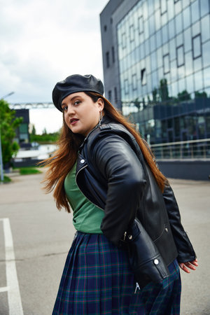 stylish woman with plus size body walking in leather jacket with black beret, plaid skirt and greet t-shirt near blurred modern building on urban street outdoors, body positive, looking at cameraの写真素材