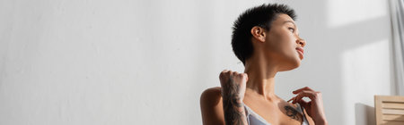young and sexy tattooed woman with short brunette hair touching straps of silk bra while looking...