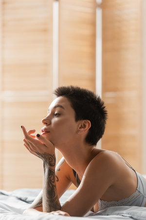 young, charming and dreamy woman with short brunette hair and sexy tattooed body looking away while laying in bra on white bedding near room divider on blurred background, boudoir photographyの写真素材