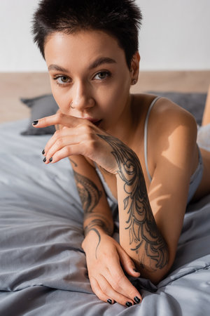 young, charming and provocative woman in lingerie, with short brunette hair and sexy tattooed body holding hand near face and looking at camera on grey bedding at homeの写真素材