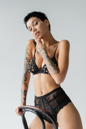 sexy and flirtatious woman with tattooed body and short brunette hair, wearing black bra with pearl beads and lace panties while looking at camera near chair on grey backgroundの写真素材