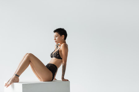 full length of young, sexy and tattooed woman with short brunette hair sitting on white cube in black bra with pearl beads and lace panties on grey background, erotic photographyの写真素材