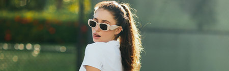flirty and seductive young woman with brunette hair in ponytail and sunglasses standing in white polo shirt and sticking out tongue on tennis court with blurred background, banner, Miami, Floridaの写真素材