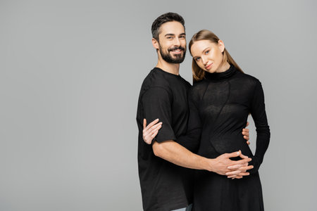 Positive and bearded husband in t-shirt looking at camera while hugging stylish pregnant wife in...