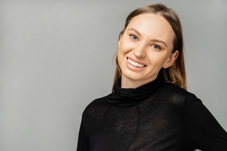 Portrait of cheerful fair haired woman with natural makeup wearing stylish black dress and looking at camera while standing isolated on grey with copy spaceの写真素材