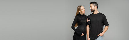 Stylish and pregnant woman in black dress touching arm of smiling bearded husband and standing...