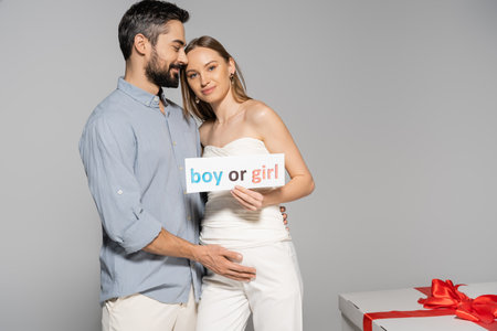 Smiling pregnant woman holding card with boy or girl lettering near stylish husband and big gift box during gender reveal surprise party isolated on grey, expecting parents conceptの写真素材