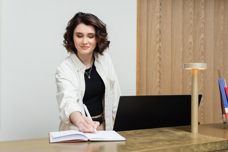 friendly concierge in stylish casual clothes, with wavy brunette hair writing in notebook near...