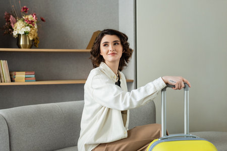 young and happy woman with wavy brunette hair and tattoo, in stylish casual clothes sitting on couch near yellow travel bag, books and vase with flowers on shelves in lobby of modern hotelの写真素材