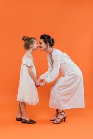 summer trends, young mother holding hands with happy preteen daughter and standing on orange background, white sun dresses, togetherness, fashion and style concept, bonding and love, nose to noseの写真素材