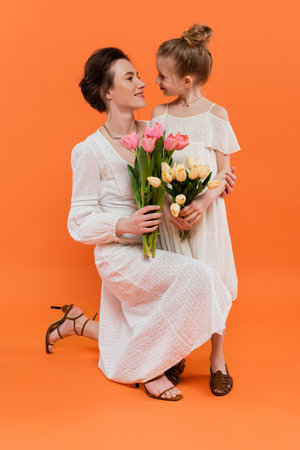 happy mother and daughter with flowers, young woman and girl holding tulips and posing on orange background, summer fashion, sun dresses, female bondingの写真素材