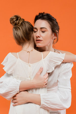 motherly love, caring mother hugging daughter on orange background, closed eyes, white sun dresses,...