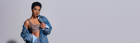 Modern short haired african american woman in top with animal print and denim clothes looking at camera and standing on grey background, denim fashion concept, bannerの写真素材