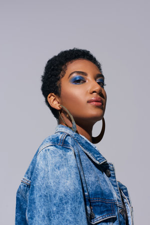 Low angle view of modern african american woman with vivid makeup and golden earrings looking at camera while posing in denim jacket isolated on grey, denim fashion conceptの写真素材