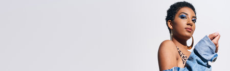 Low angle view of trendy young african amercan model with golden earrings looking at camera and posing in denim jacket while standing isolated on grey, denim fashion concept, bannerの写真素材