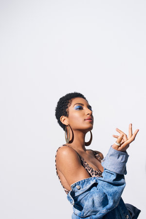 Young short haired african american woman with bold makeup posing in top with animal print and denim jacket while looking away isolated on grey, denim fashion conceptの写真素材
