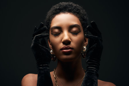 Portrait of elegant and short haired african american woman with evening makeup, golden chains, earrings and gloves posing with closed eyes isolated on black, high fashion and evening lookの写真素材