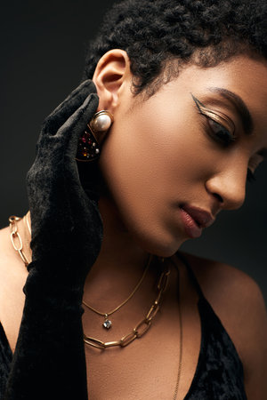 Portrait of fashionable african american woman with evening makeup and golden necklaces wearing glove and touching earring isolated on black, high fashion and evening lookの写真素材