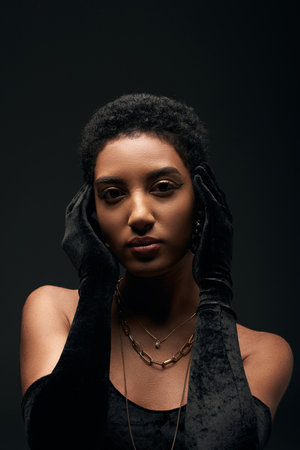 Portrait of fashionable and short haired african american woman with makeup and golden necklaces wearing gloves and dress while standing isolated on black, high fashion and evening lookの写真素材