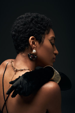 Side view of fashionable african american model in glove and modern golden chains and earring touching back and standing isolated on black, high fashion and evening lookの写真素材