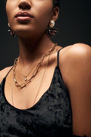 Cropped view of young and elegant african american model in dress and golden chains and earrings standing isolated on black under lighting, high fashion and evening look, details, close upの写真素材