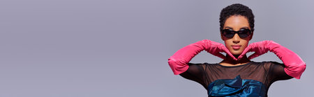 Trendy young african american model in sunglasses, pink gloves and cocktail dress posing isolated on grey with copy space, modern generation z fashion concept, bannerの写真素材