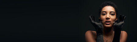 Fashionable african american model with evening makeup wearing gloves and holding golden chain near mouth isolated on black with lighting, high fashion and evening look, bannerの写真素材