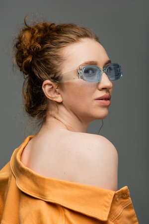 Modern red haired woman in blue sunglasses and orange denim jacket with naked shoulder looking away...