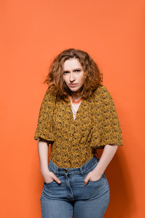 Serious young red haired model in blouse with abstract print and jeans holing hands in pockets and...