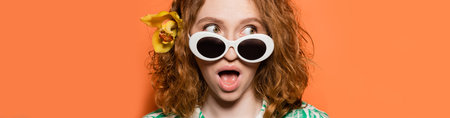 Young and shocked redhead woman with orchid flower and stylish sunglasses looking away while...