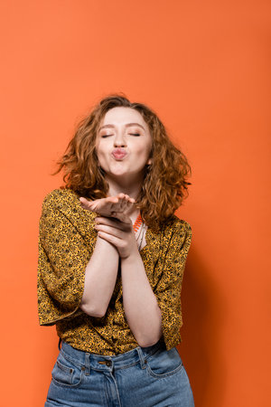 Young and stylish red haired woman in yellow blouse with abstract pattern and jeans blowing air kiss at camera on orange background, stylish casual outfit and summer vibes concept, Youth Cultureの写真素材