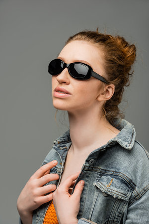 Red haired and freckled young woman in stylish sunglasses posing in denim jacket and top with...