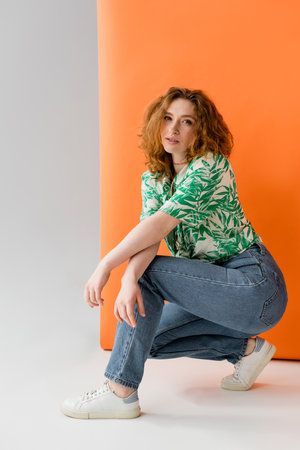 Full length of stylish redhead woman in blouse with floral pattern and jeans looking at camera while posing on orange and grey background, trendy casual summer outfit concept, Youth Cultureの写真素材