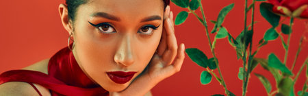 portrait of youthful and alluring asian woman with bold makeup and expressive gaze holding hand near face and looking at camera near flowers on red background, trendy spring, generation z, bannerの写真素材