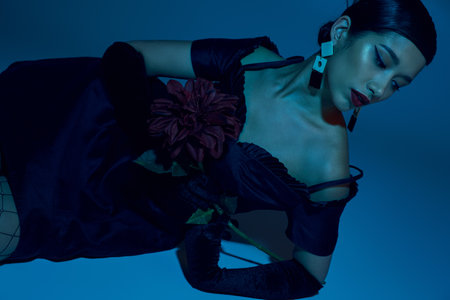 young and sensual asian woman in trendy earrings, black elegant dress and long gloves laying with...