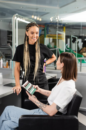 happy beauty worker standing near female client, holding hair color palette, tattooed woman choosing hair color, customer in salon, hair extension, hair trends, tattoo translation: loveの写真素材