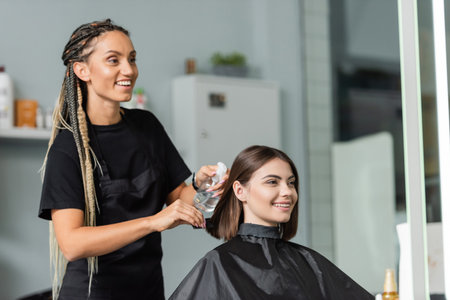 hairstylist spraying hair of happy woman, hairdresser with braids holding spray bottle near female client with short brunette hair in salon, hair treatment, hair make over, hairdoの写真素材