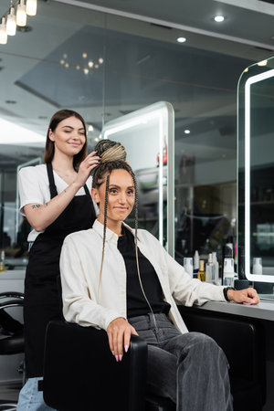 hair make over, cheerful hair stylist doing hair bun to female client with braids, cheerful women, client satisfaction, customer in salon, beauty service, salon experience, feminine, tattooedの写真素材