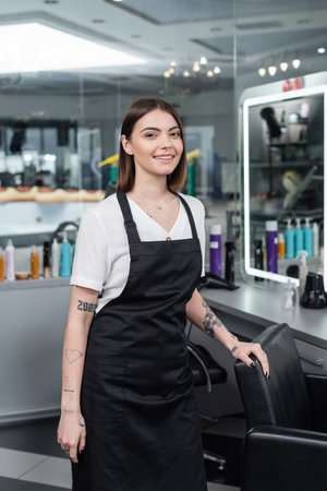professional headshots, tattooed beauty worker in apron looking at camera in beauty salon, hairdressing chair, hair stylist, hair coloring, salon job, beauty salon work, hair trendsの写真素材