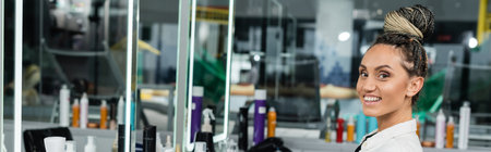cheerful client in beauty salon, happy woman with hair bun looking at camera, customer satisfaction, hair salon, hairstyle, female client with braids, beauty service, mirror, bannerの写真素材