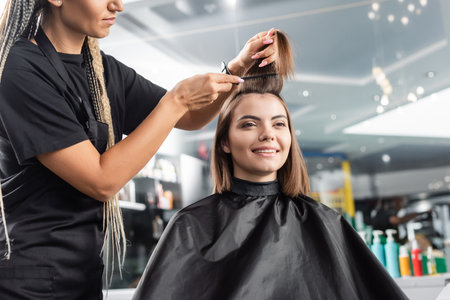 beauty profession, hairdo, hairdresser with braids brushing short brunette hair of happy woman, haircut, professional, beauty salon work, haircut, hairdressing cape, client satisfactionの写真素材